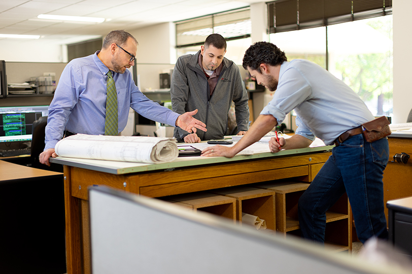 Three employees discussing blueprints that are unrolled on drafting table