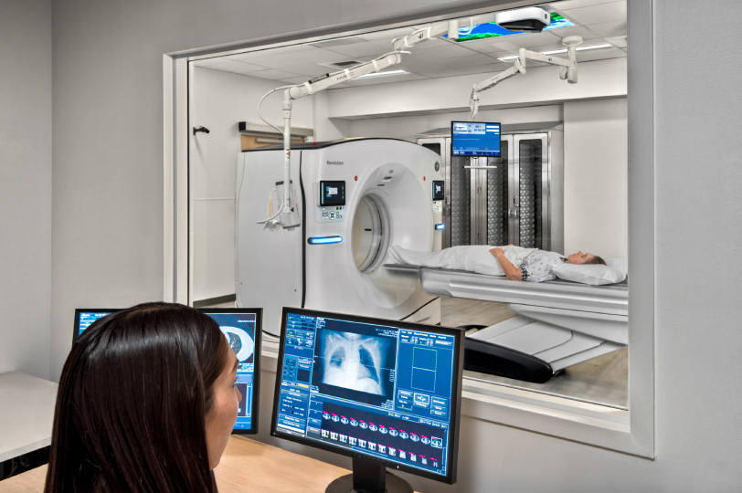Medical technician operating CT scanner from control room while patient undergoes scan